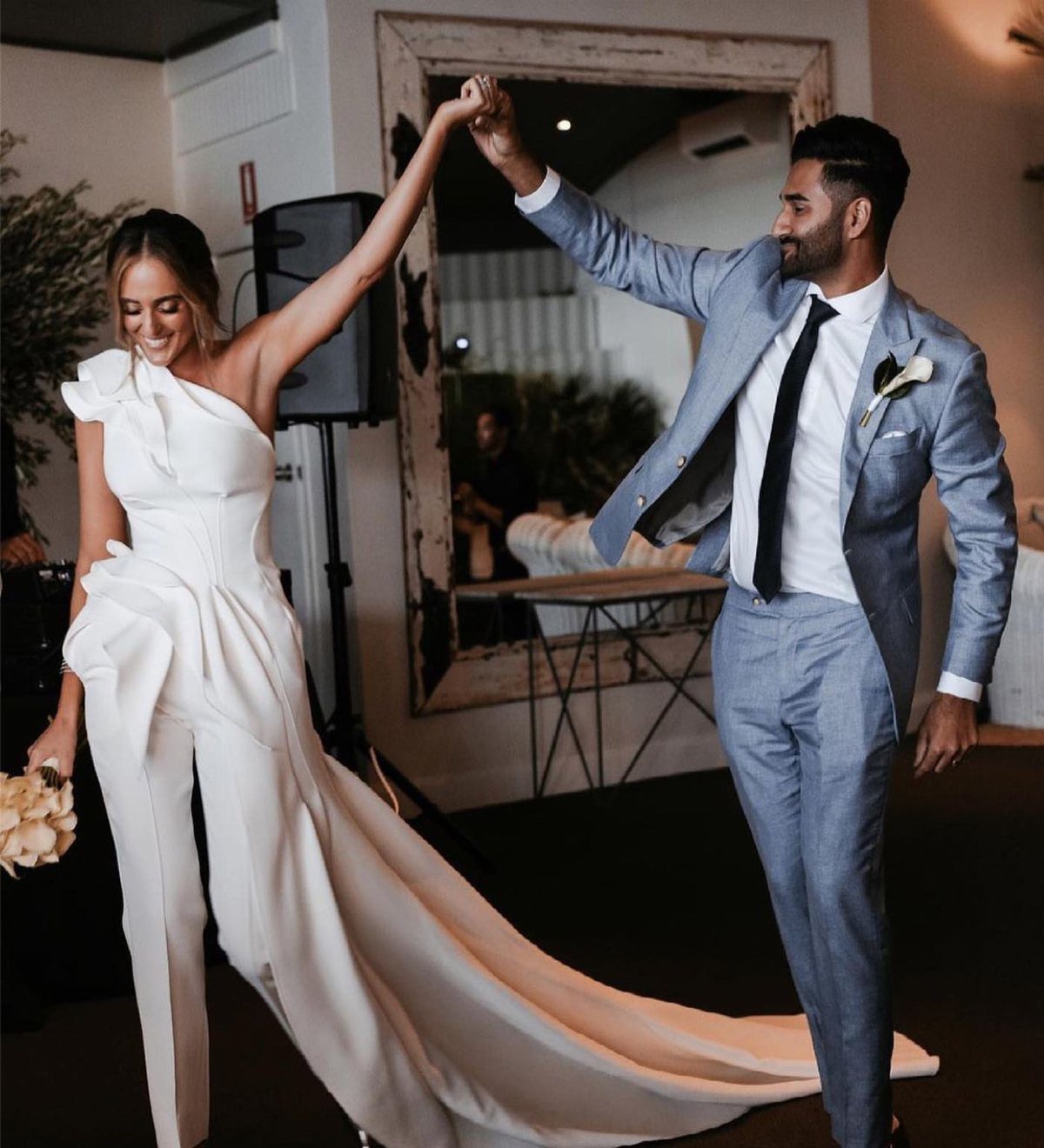 Woman in white bridal jumpsuit and man in blue suit dancing together indoors.