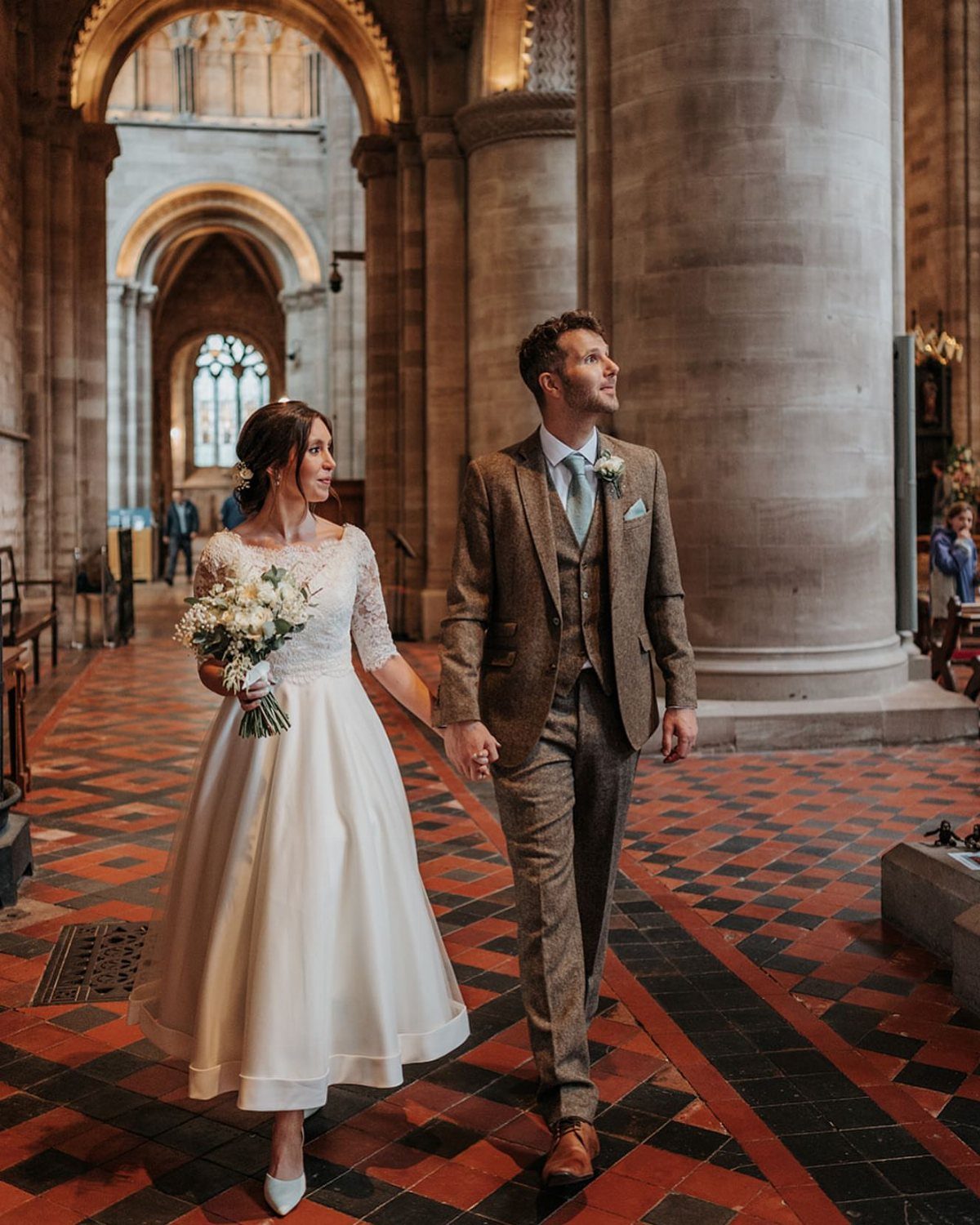 Bride in white tea-length gown and groom in brown tweed suit walking in cathedral.