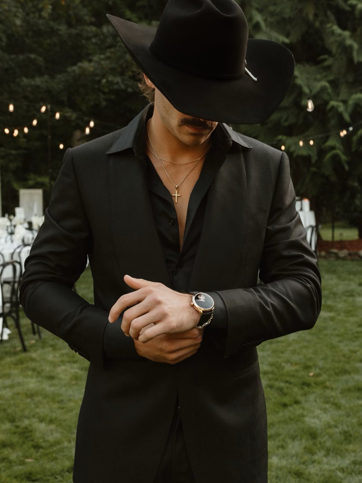 Man in black cowboy hat and blazer adjusting his watch at outdoor event