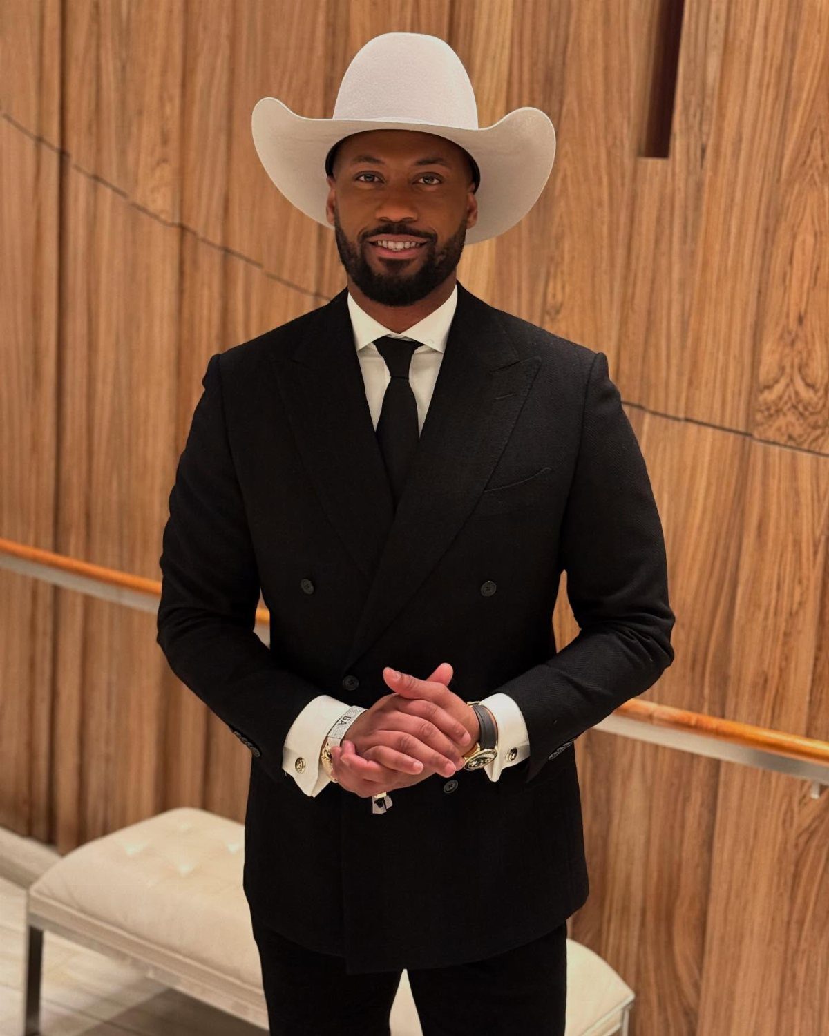 Man in a black double-breasted suit and white cowboy hat posing against wood paneling.