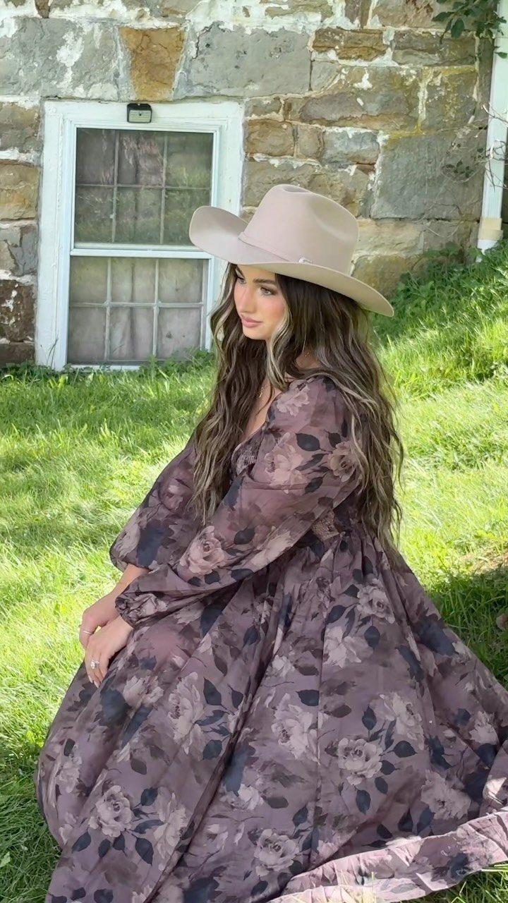 Woman in a purple floral maxi dress and tan cowboy hat sitting on grass.