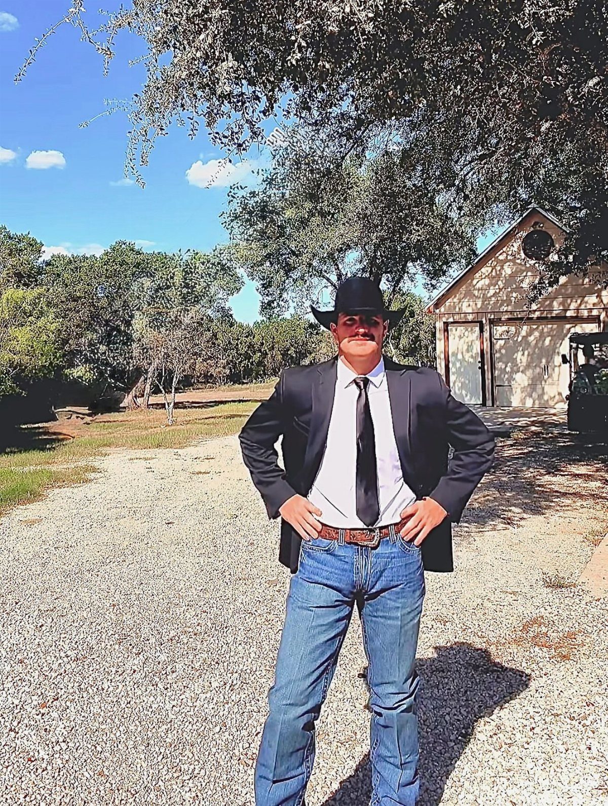 Man in black cowboy hat and suit coat posing on a gravel path.