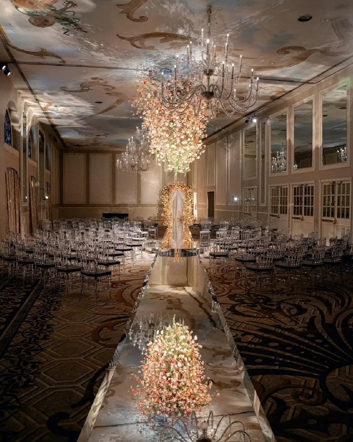 Opulent ballroom wedding setup with mirrored aisle, clear chairs, and grand floral arrangements.