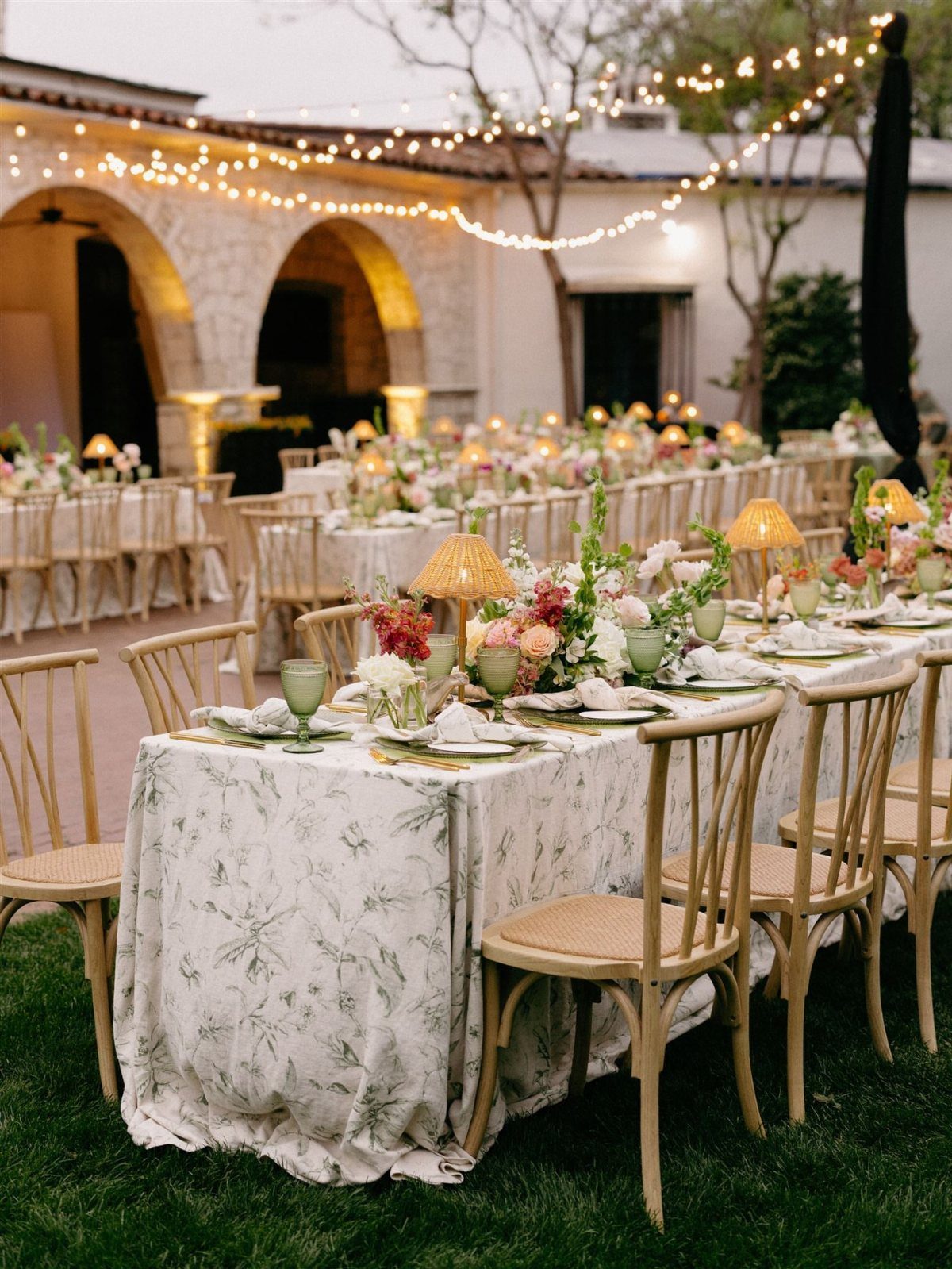 Outdoor wedding reception tables with floral linens and string lights in a garden setting.