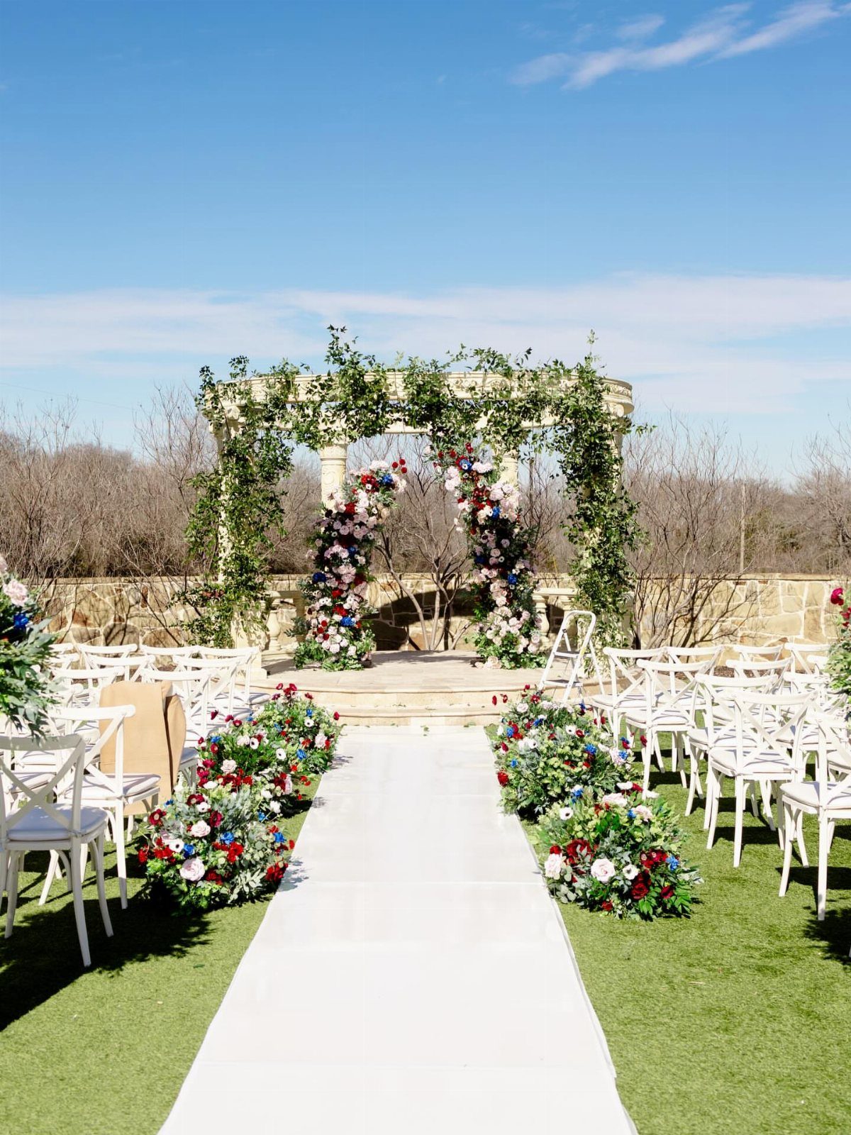 Outdoor wedding ceremony setup with white aisle, floral arch, and white chairs on grass.