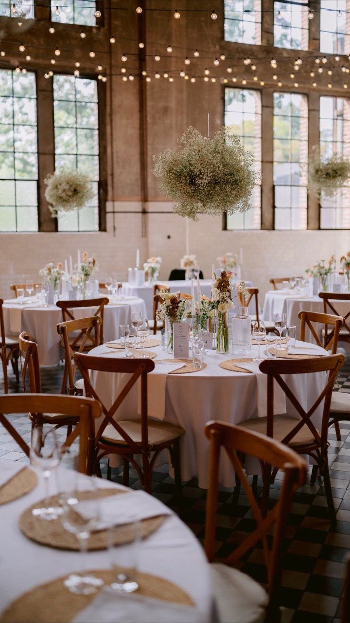 Industrial wedding venue with white tables, wooden chairs, and large windows with string lights.