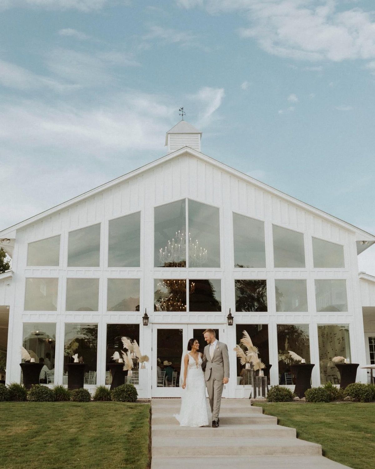 Bride and groom walking before a large white modern barn wedding venue.