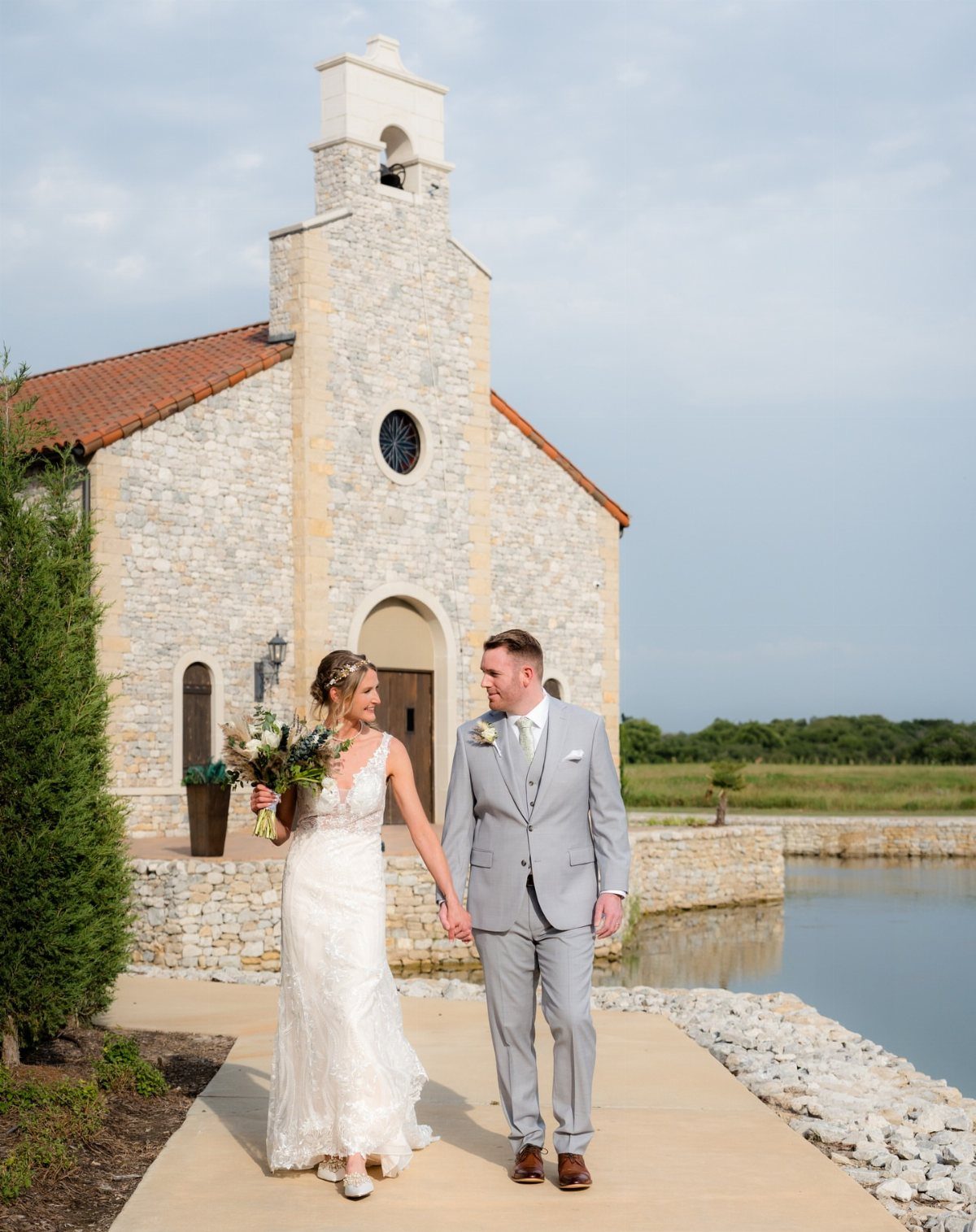 Bride and groom walking hand-in-hand in front of a stone wedding chapel by a pond.