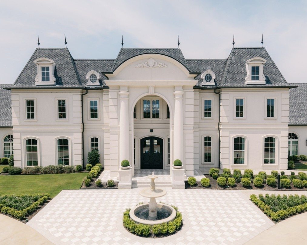 White French-inspired estate with a grey roof and a tiered stone courtyard fountain.