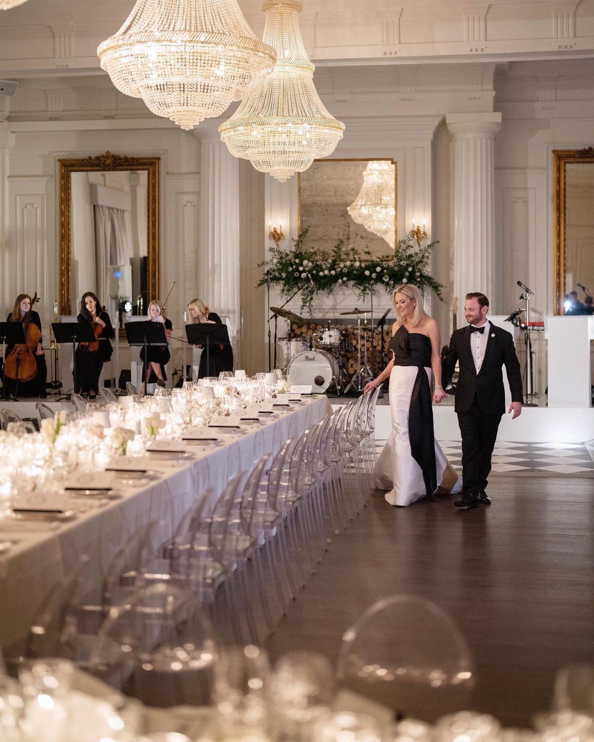 Couple walking past a long wedding table under large crystal chandeliers in a ballroom