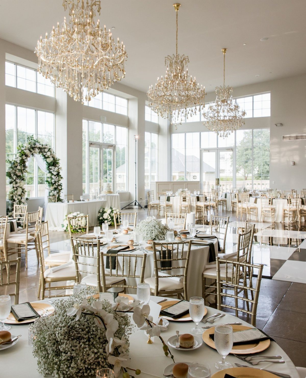 Grand event space with crystal chandeliers, gold chairs, and white floral table settings.