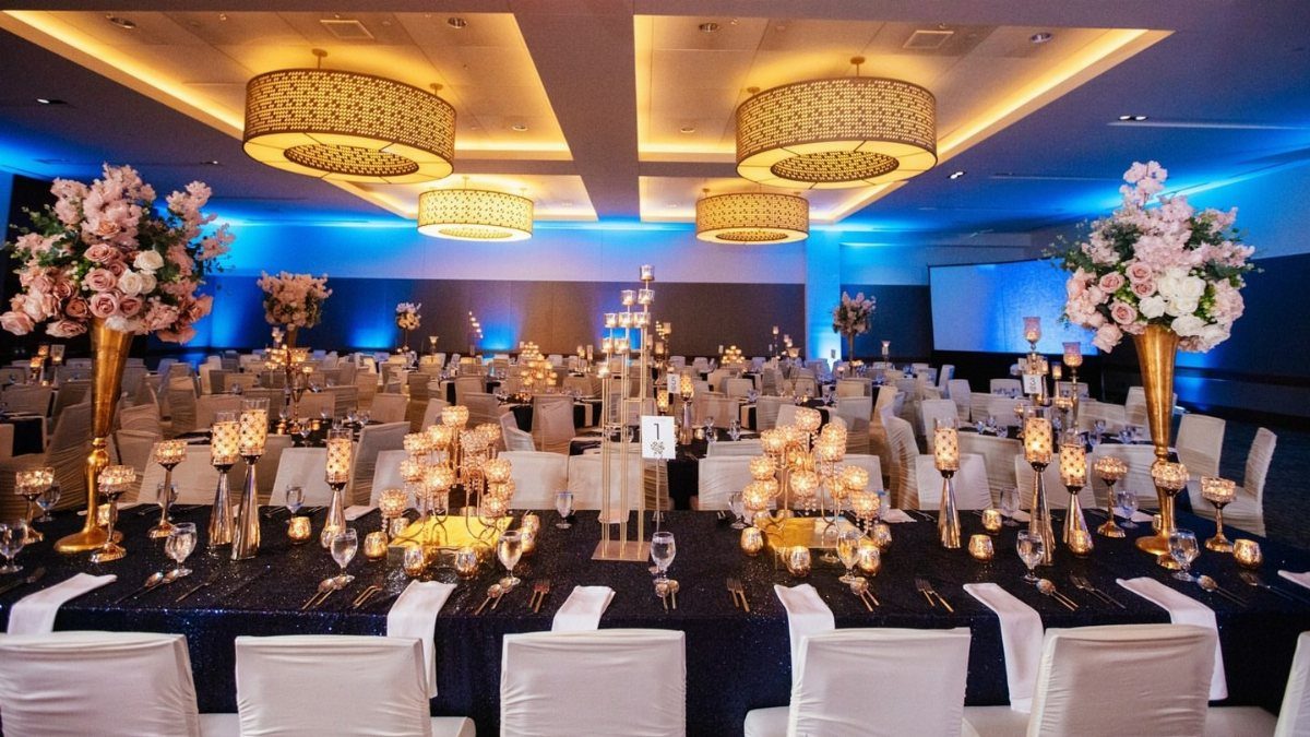 Elegant hotel ballroom with blue sequin tables, gold chandeliers, and pink floral arrangements.