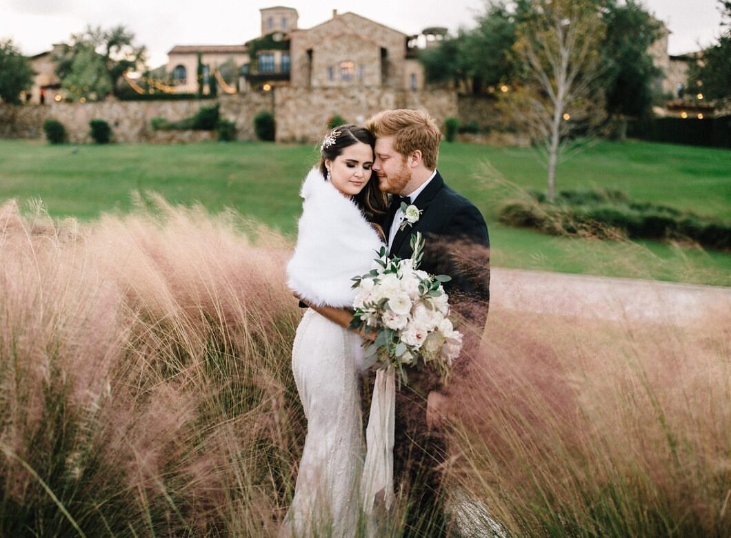 Bride and groom embracing in tall grass before a Tuscan-style stone villa venue.