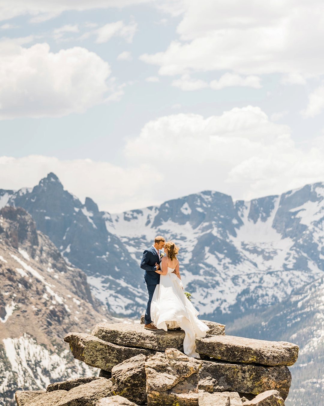 Bride and groom standing on a rocky peak overlooking snow-capped mountains.