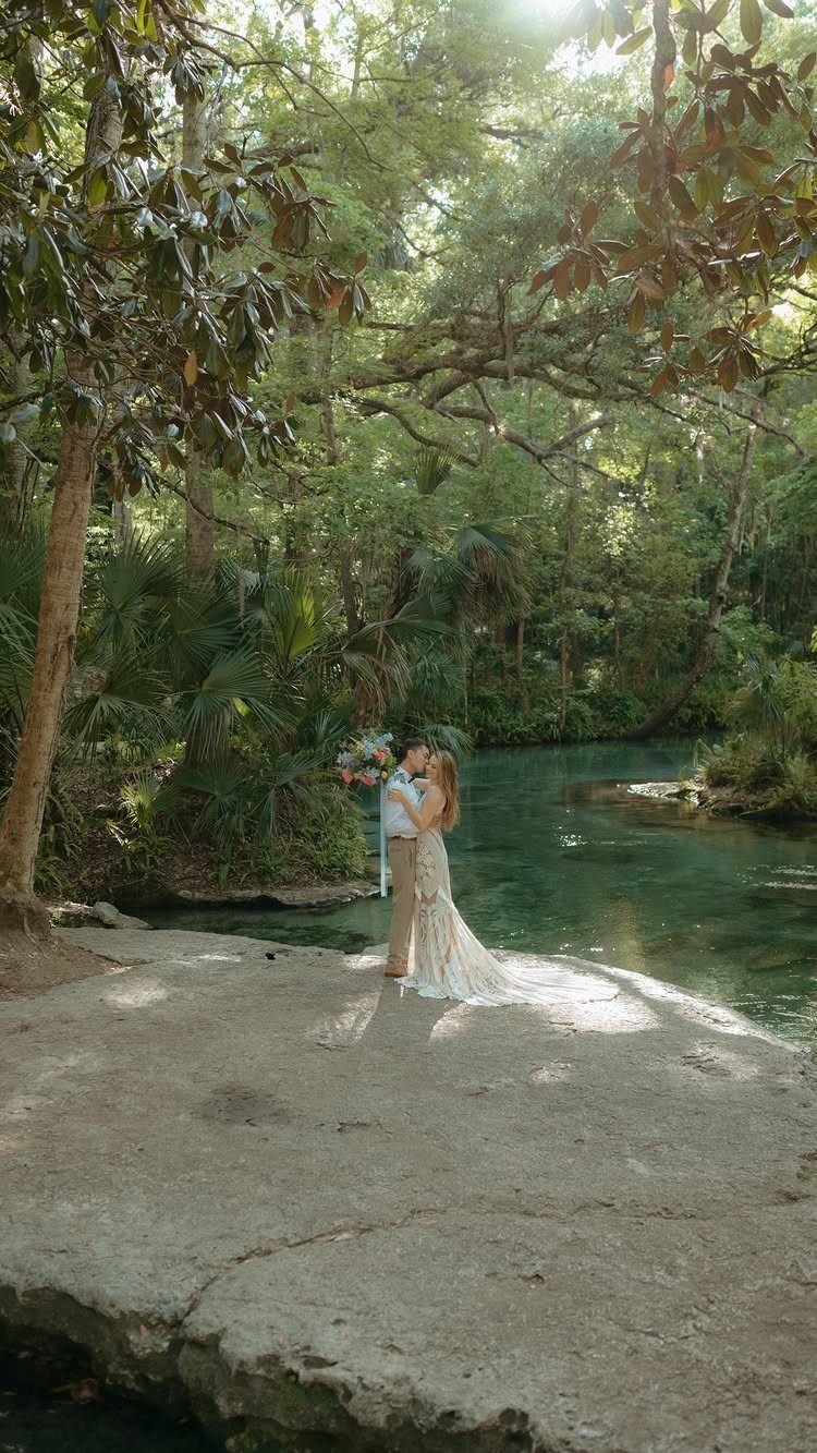 Bride and groom embracing by a turquoise river surrounded by lush green forest.