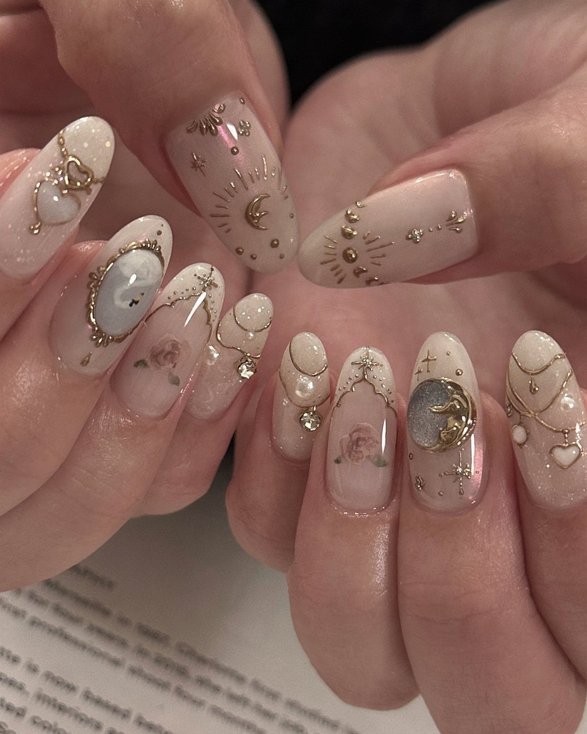 Almond nails with nude polish, gold celestial moons, stars, pearls, and floral accents.