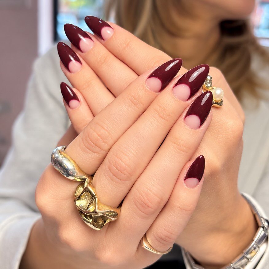 Almond-shaped nails with deep burgundy French tips and gold rings on hands.