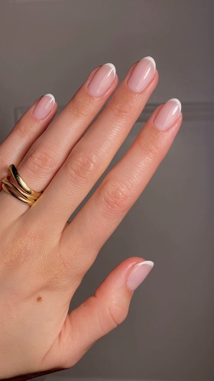 Hand with almond-shaped French manicure nails and gold rings on a grey background.