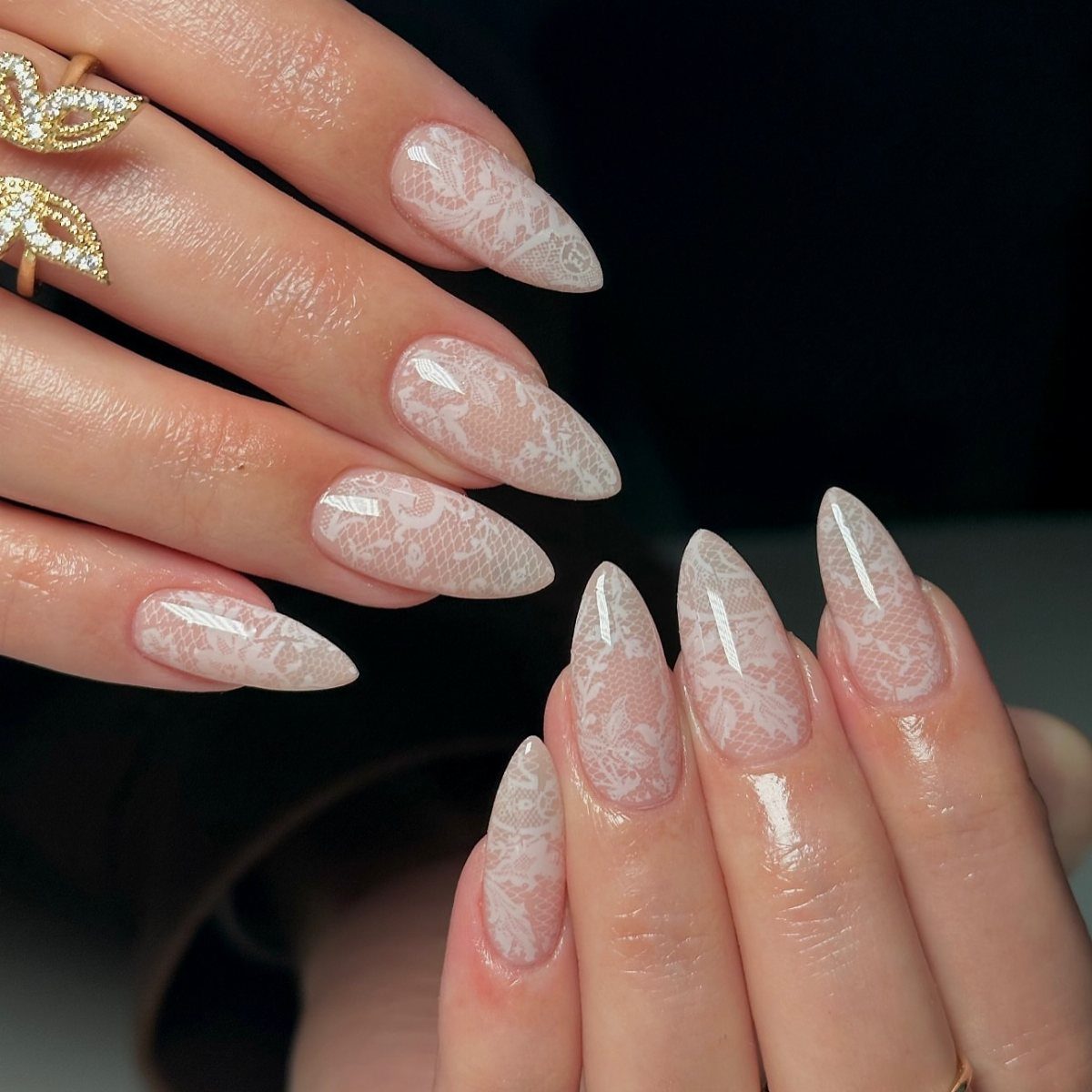 Almond-shaped nails with white lace patterns over a sheer pink base and gold ring.