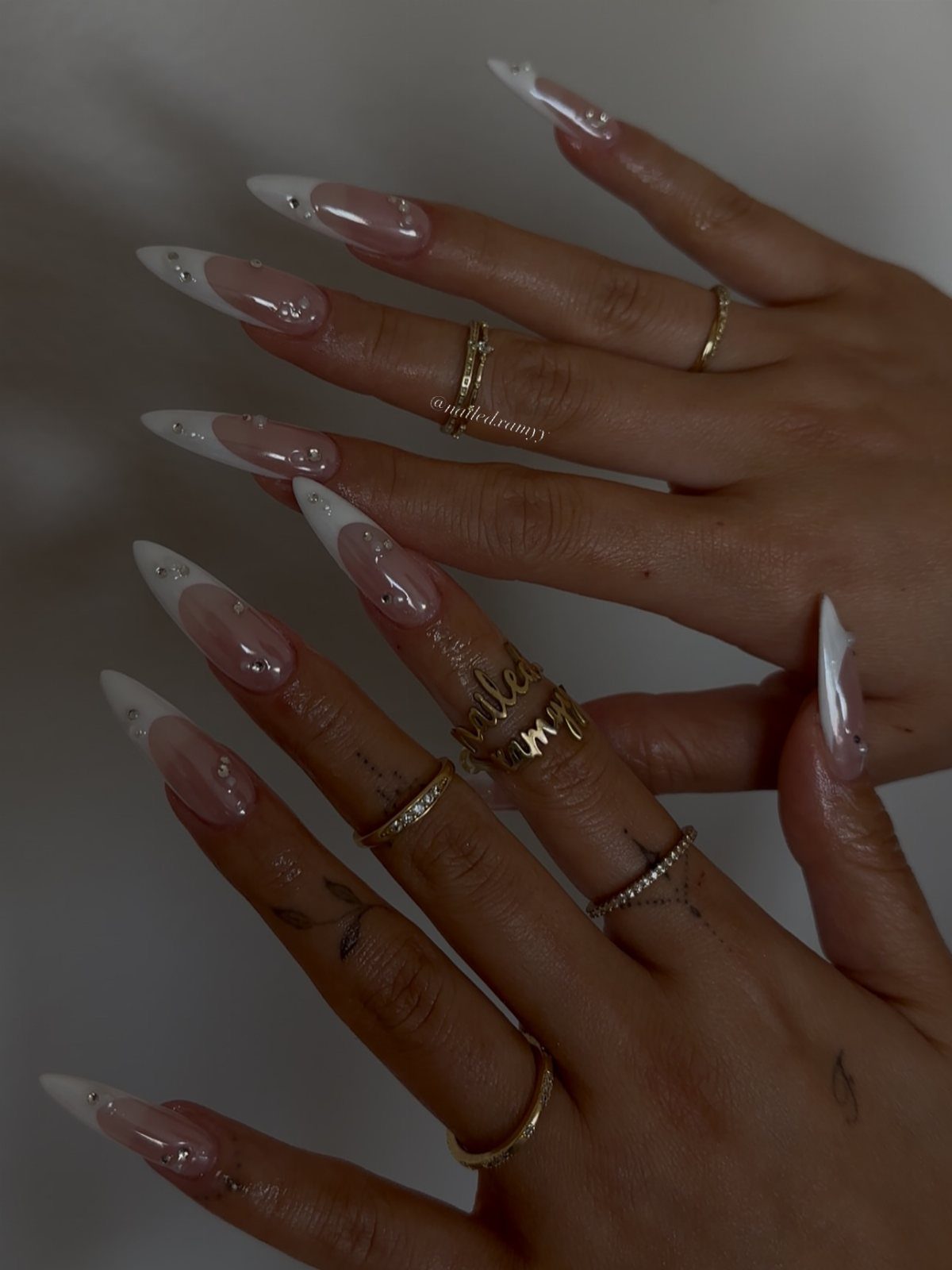 Almond-shaped French manicure with rhinestones and gold rings on a person's hands.