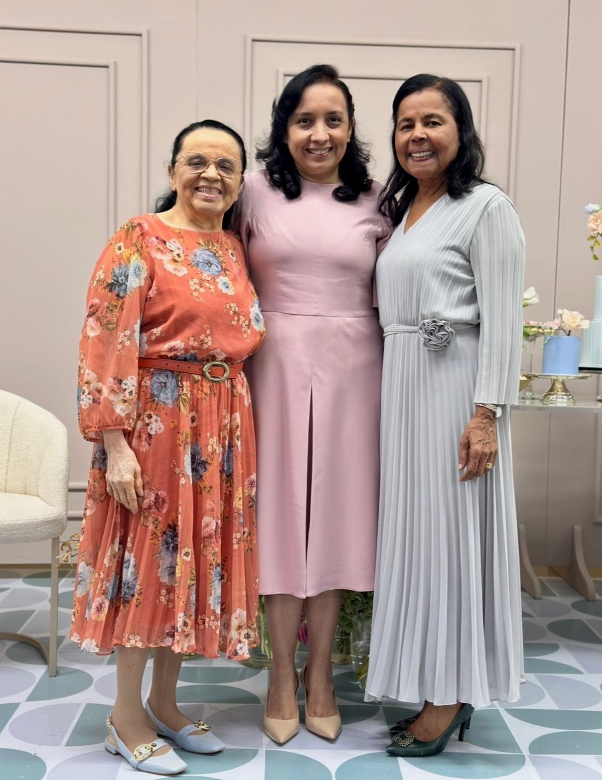 Three women wearing elegant orange floral, pink, and grey midi and tea-length dresses.