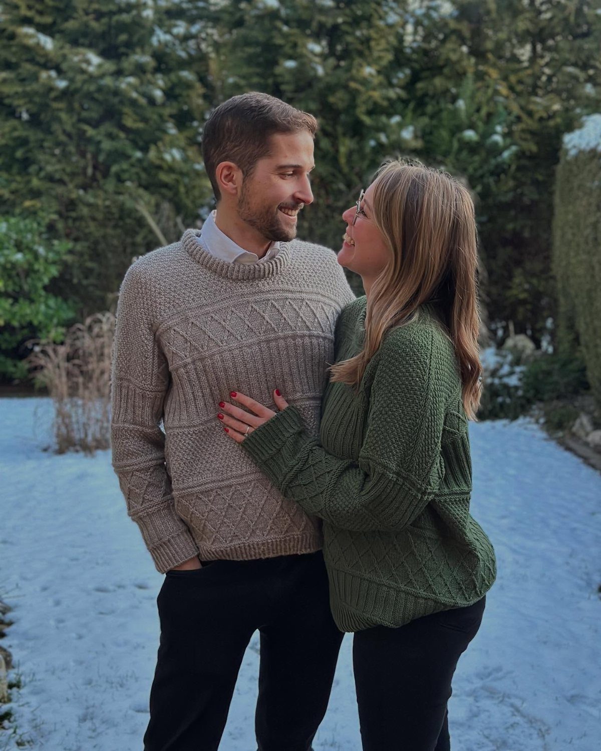 Couple wearing cable-knit sweaters in beige and olive green standing in snowy landscape