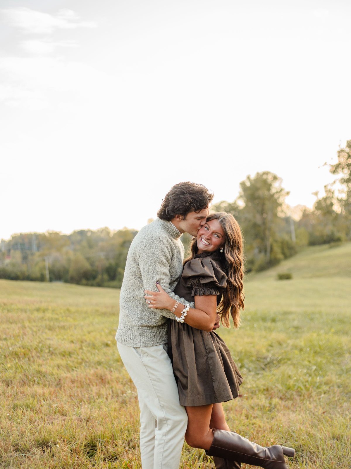 Couple in autumn outfits embracing in a grassy field with rolling hills.