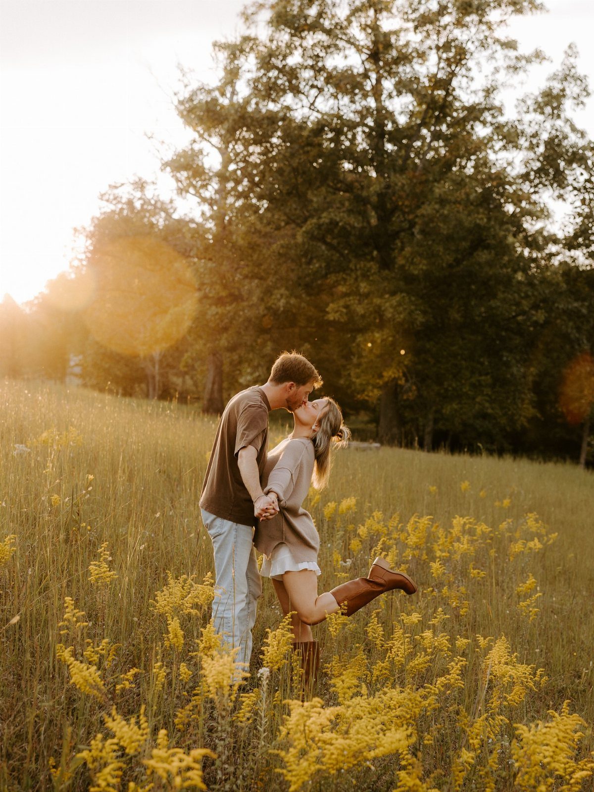 Couple kissing in a yellow wildflower field wearing autumn-toned engagement outfits.