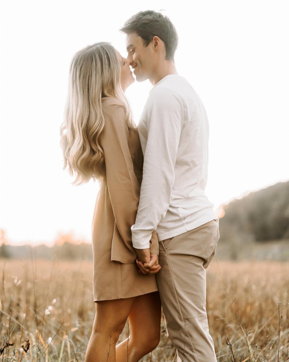 Couple kissing in a field wearing tan and white autumn engagement outfits.