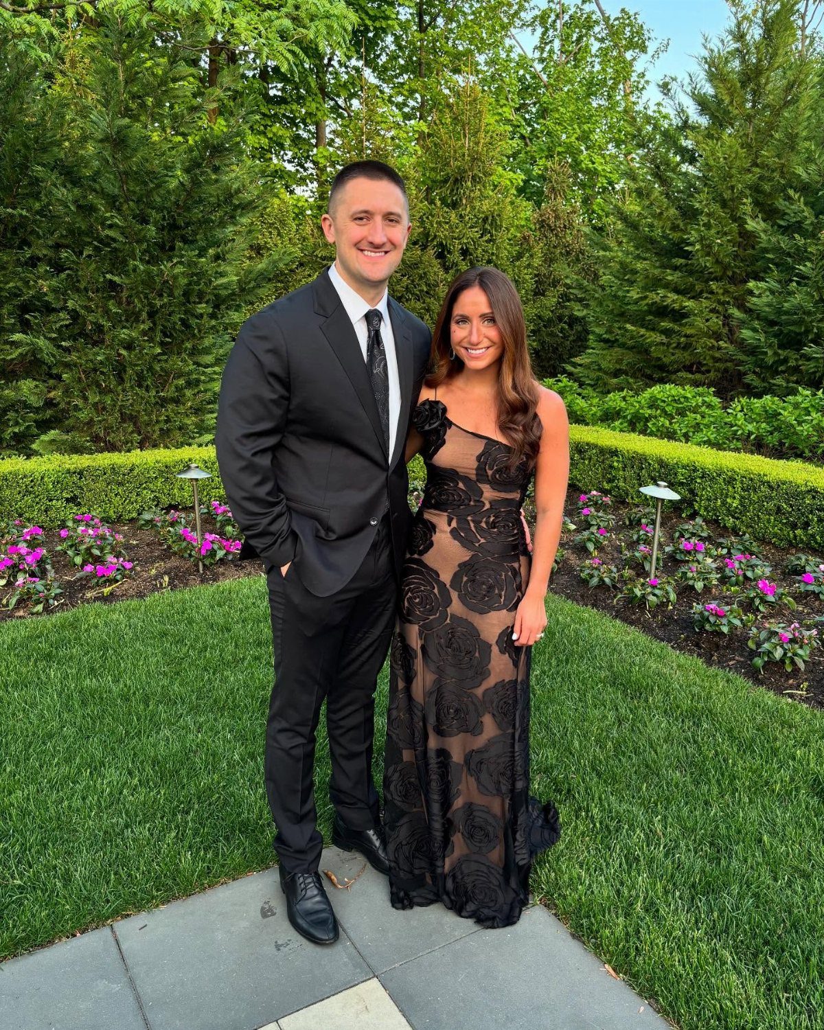 Couple in formal black tie attire posing in a green garden with flowers.