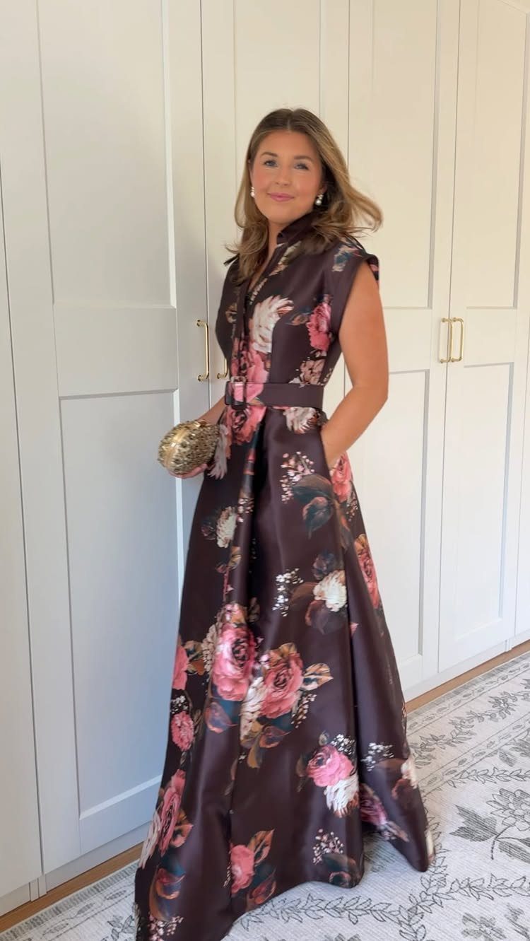 Woman in a brown floral satin gown holding a gold clutch bag indoors.