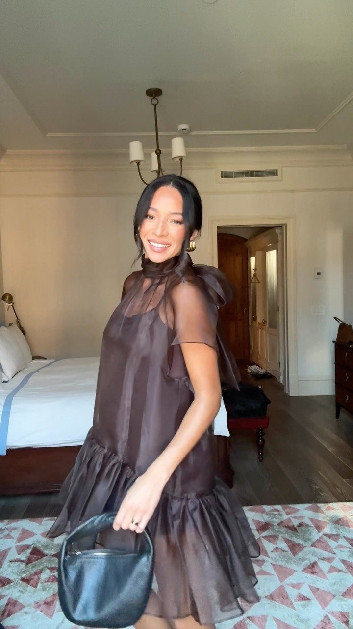 Woman in a sheer brown tiered dress and black handbag posing in a bedroom.