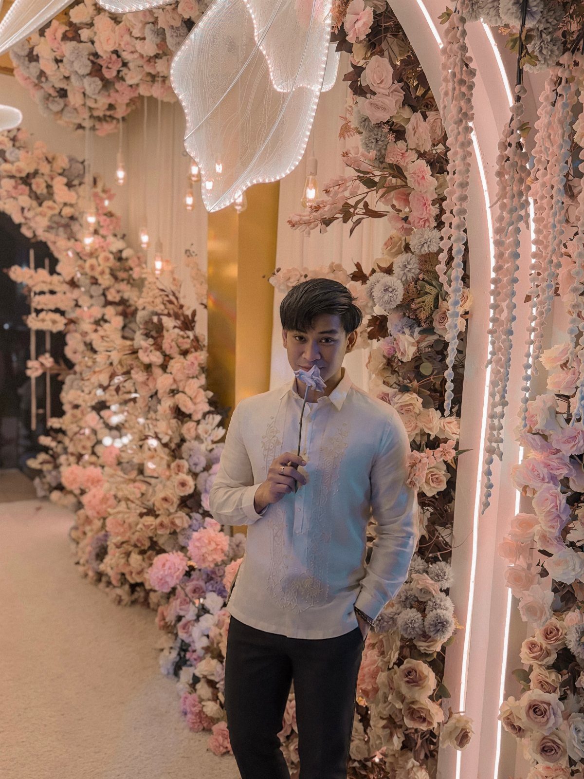 Man wearing a white embroidered Barong Tagalog standing before a floral wall.