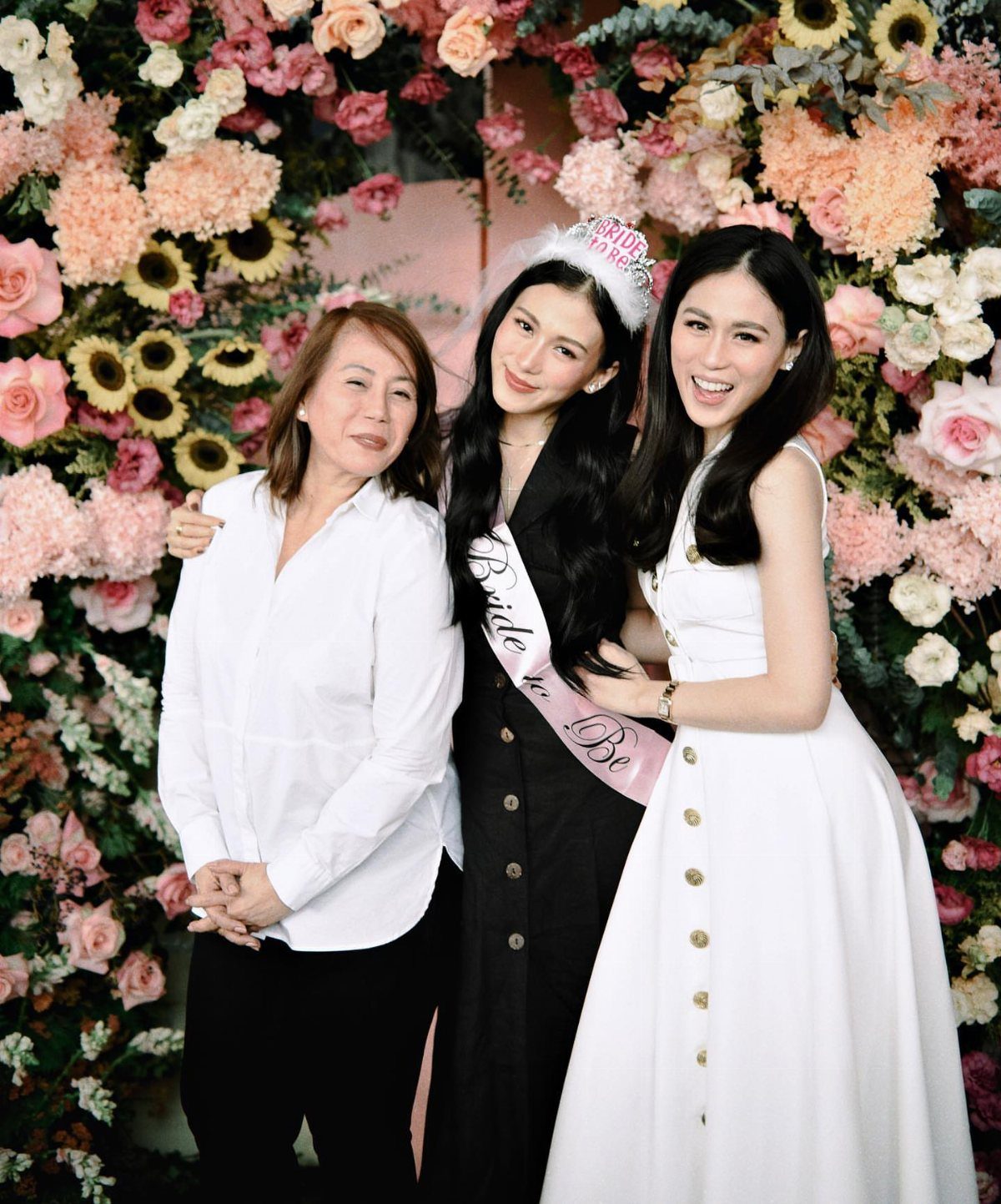 Three women smiling before a floral wall, with the bride wearing a sash.