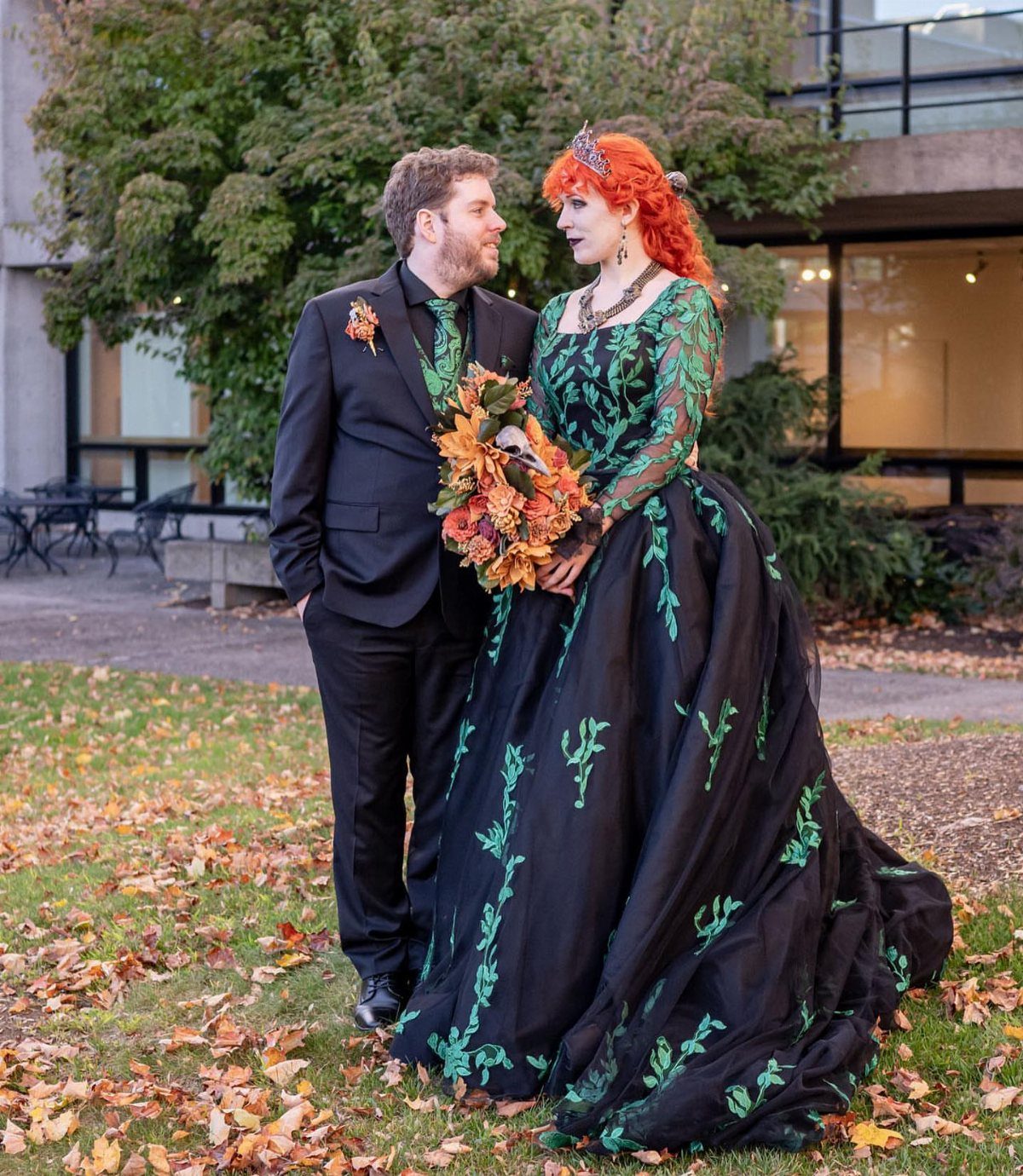 Couple in black and green wedding attire posing on autumn leaves in a park