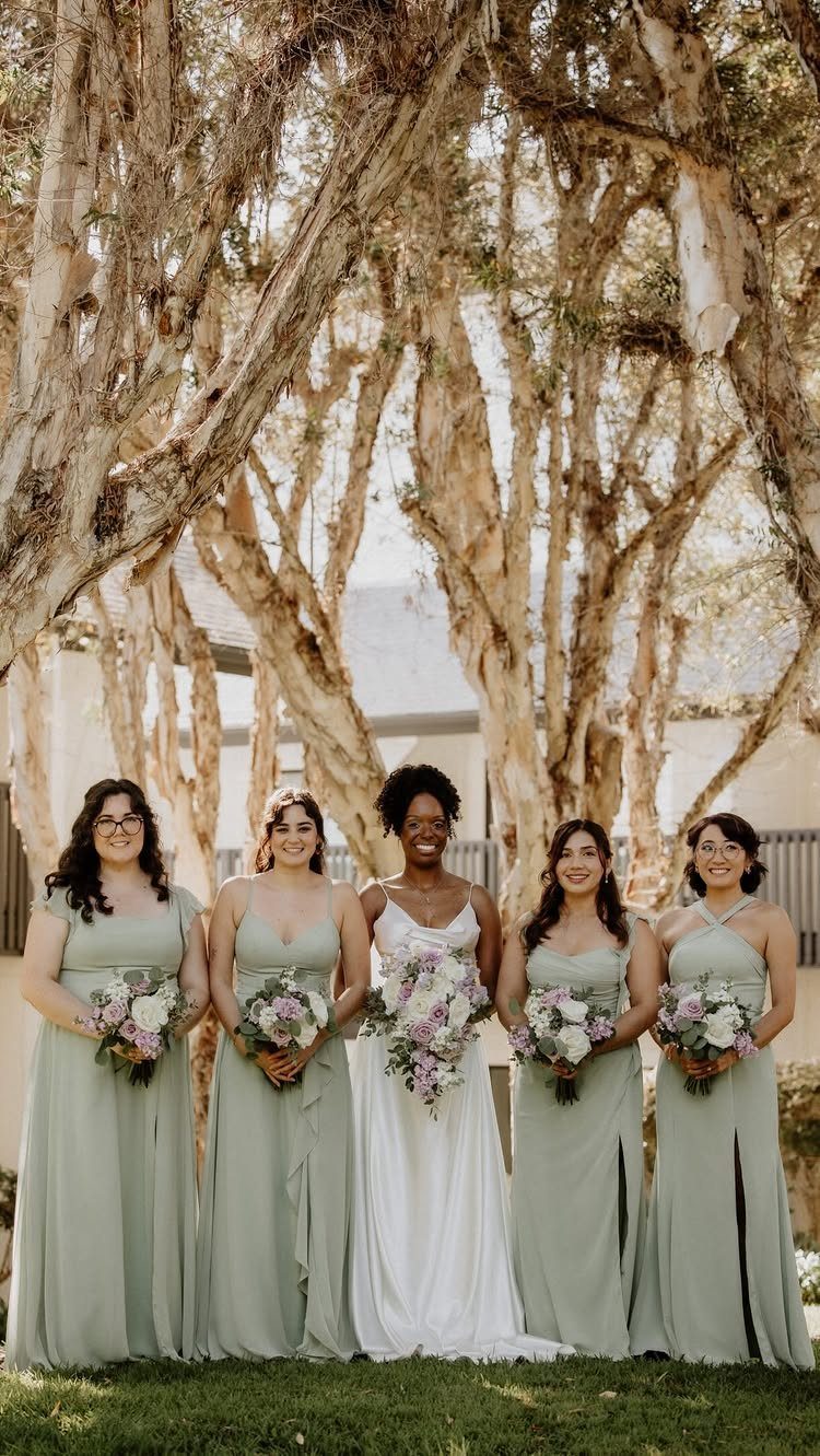 Four bridesmaids in sage green dresses and a bride in white under forest trees
