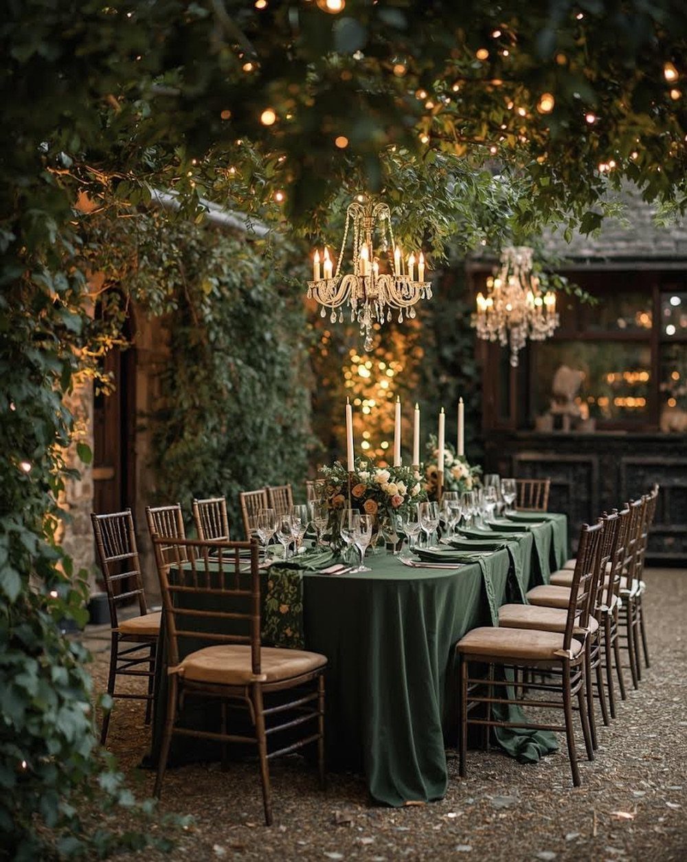 Elegant outdoor wedding table with green linens, crystal chandeliers, and twinkling overhead lights.