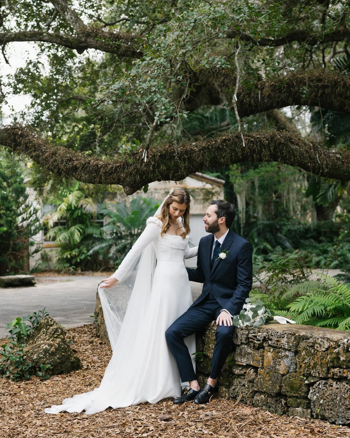 Bride in white wedding dress and groom in navy suit under a large oak tree.