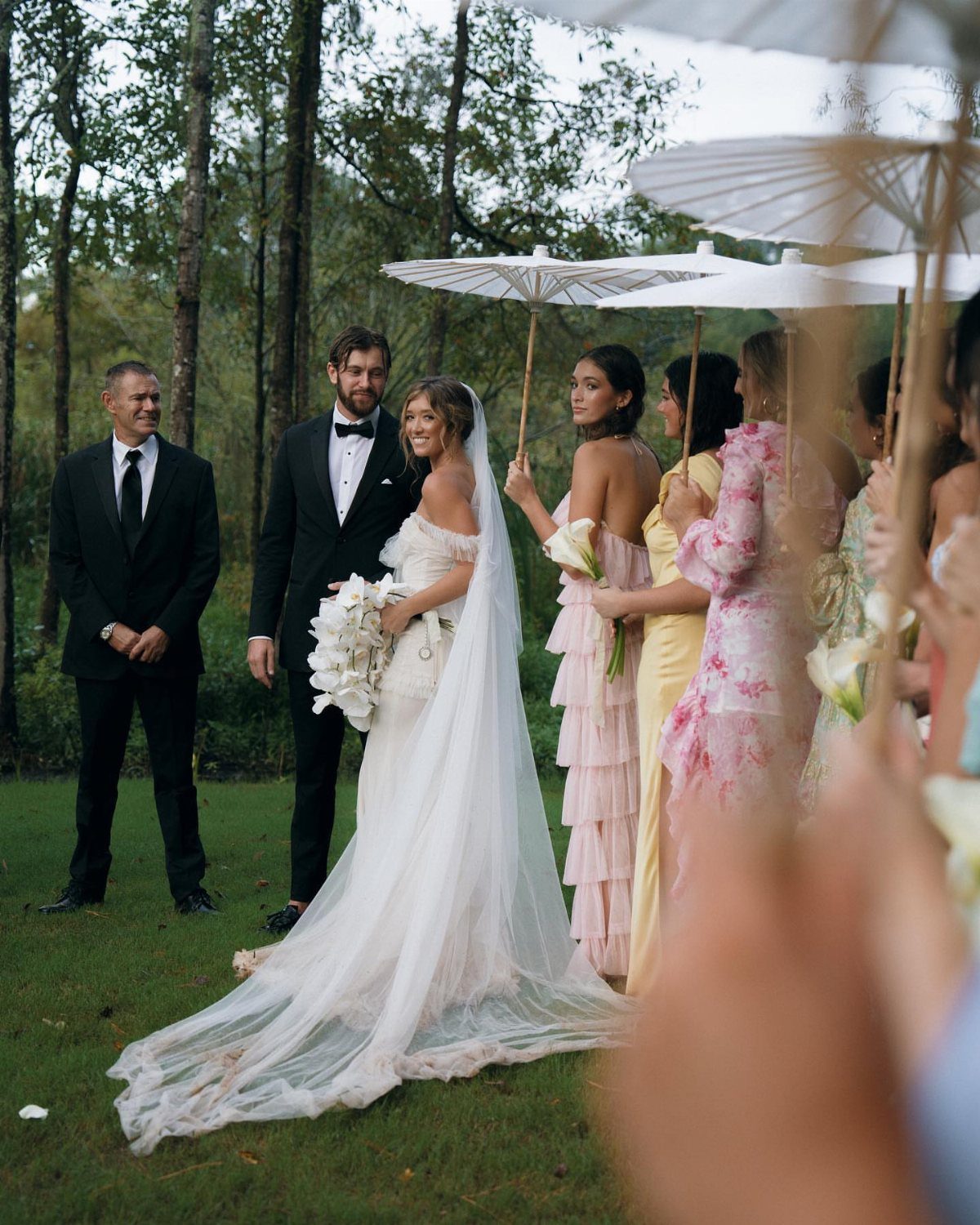Bride and groom standing with bridesmaids holding white parasols at an outdoor garden wedding.