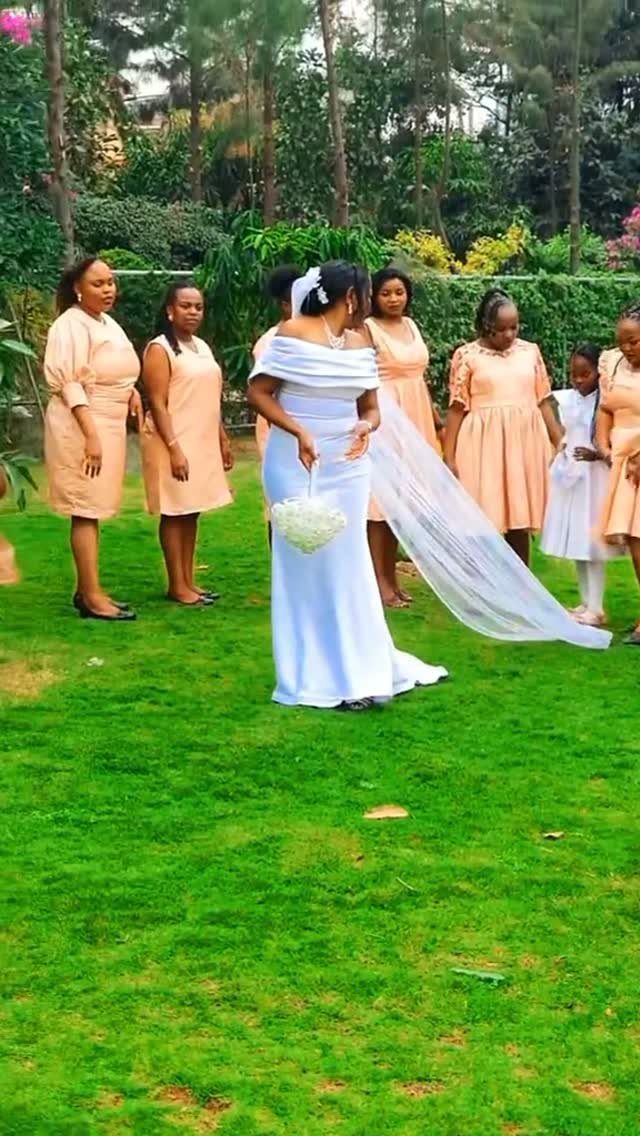 Bride in white gown and bridesmaids in peach dresses standing on a green lawn.