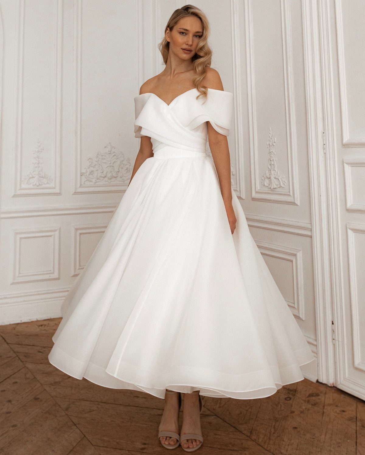Woman wearing a white off-the-shoulder tea-length wedding dress in a white paneled room.