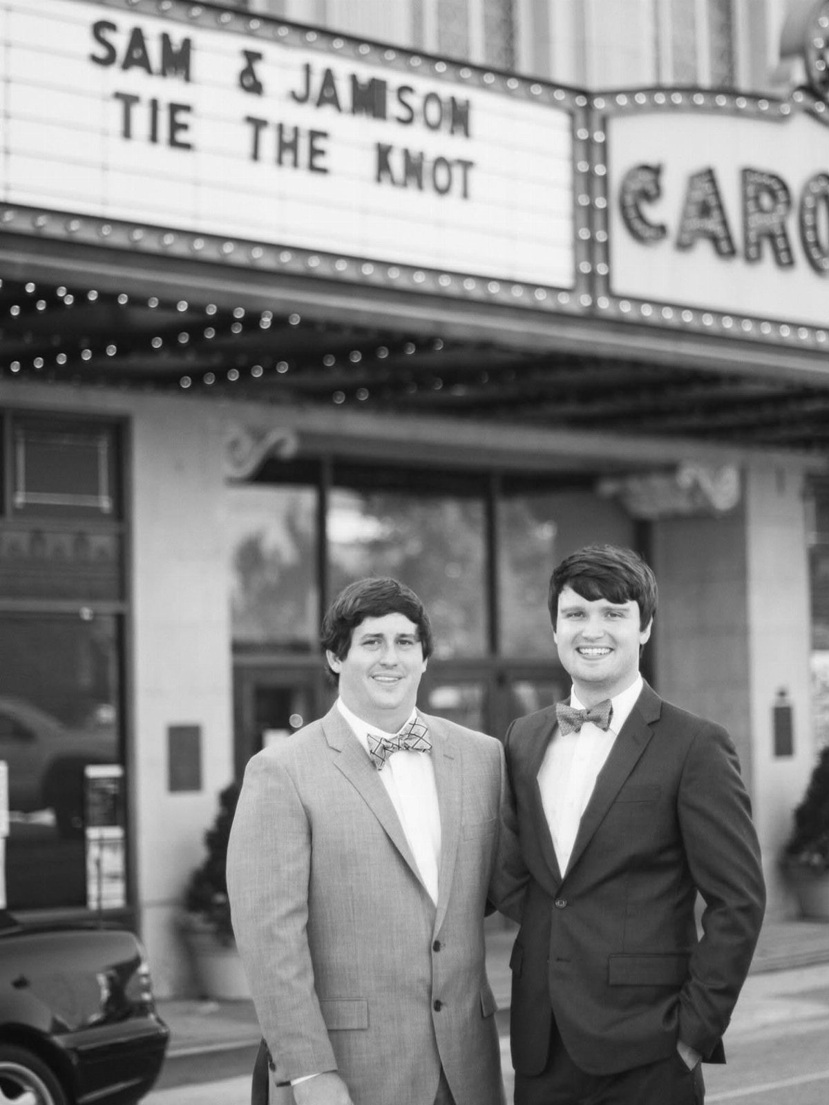 Two men wearing suits and bow ties posing before a wedding marquee.