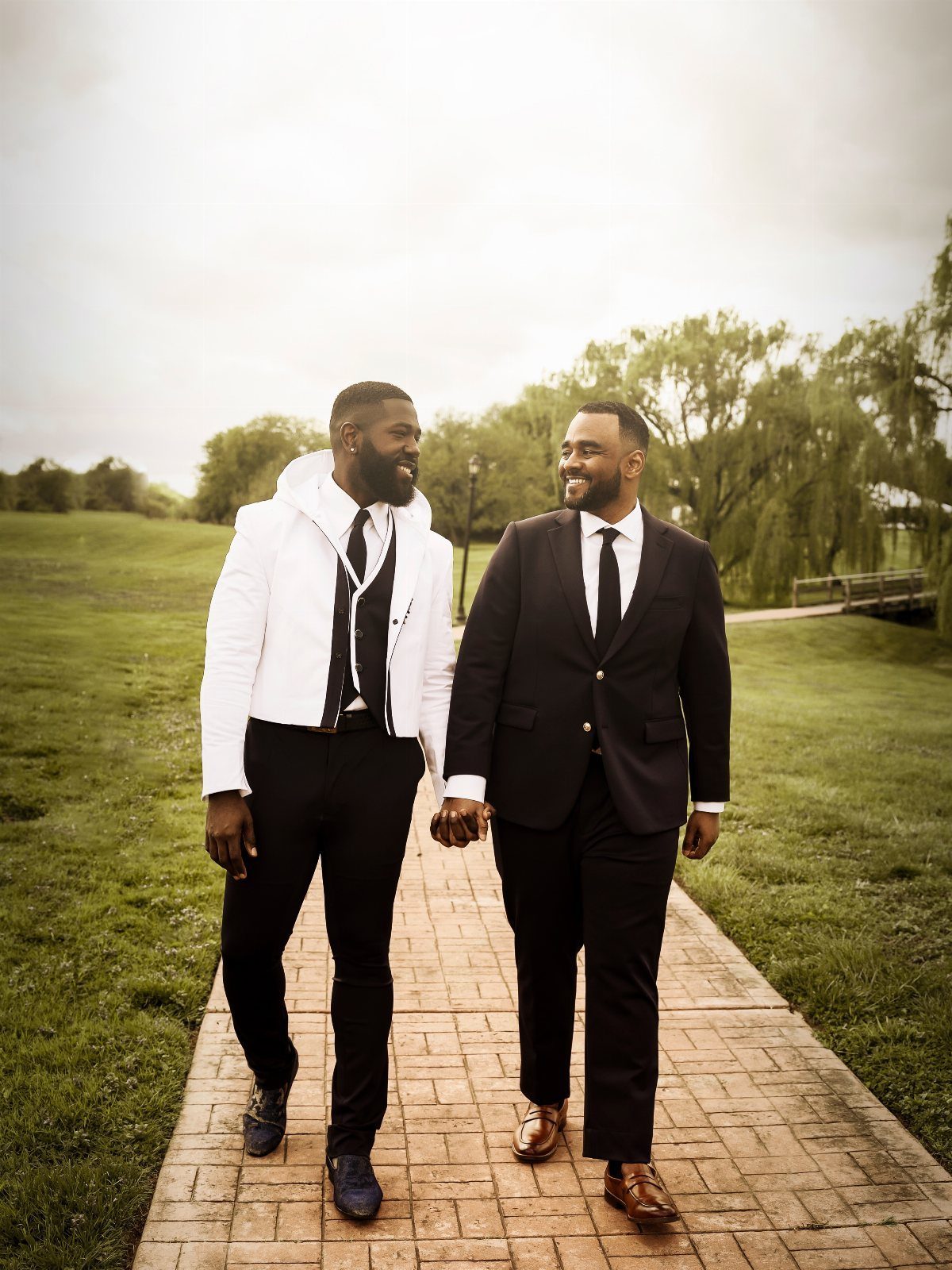 Two men in formal wedding suits holding hands while walking on a brick path.