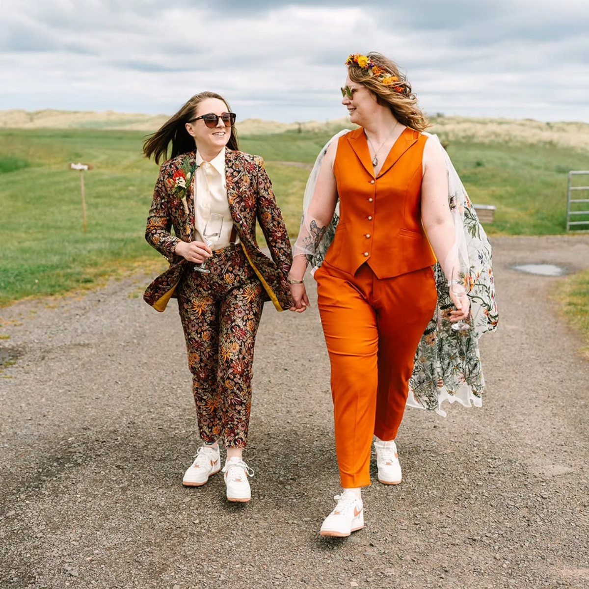 Two women in wedding suits, one floral and one orange, walking outdoors holding hands.