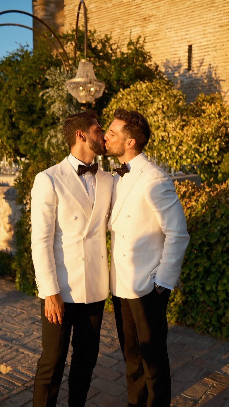 Two men in white tuxedo jackets and black trousers kissing outdoors during a wedding.