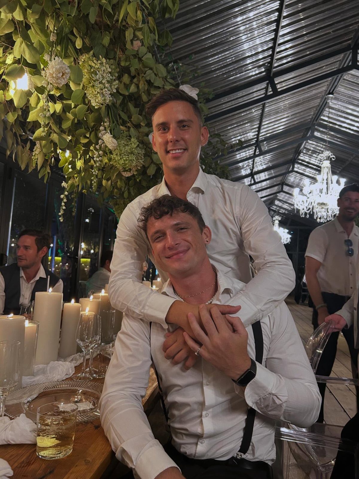 Two men wearing white dress shirts and suspenders posing at a candlelit wedding dinner.