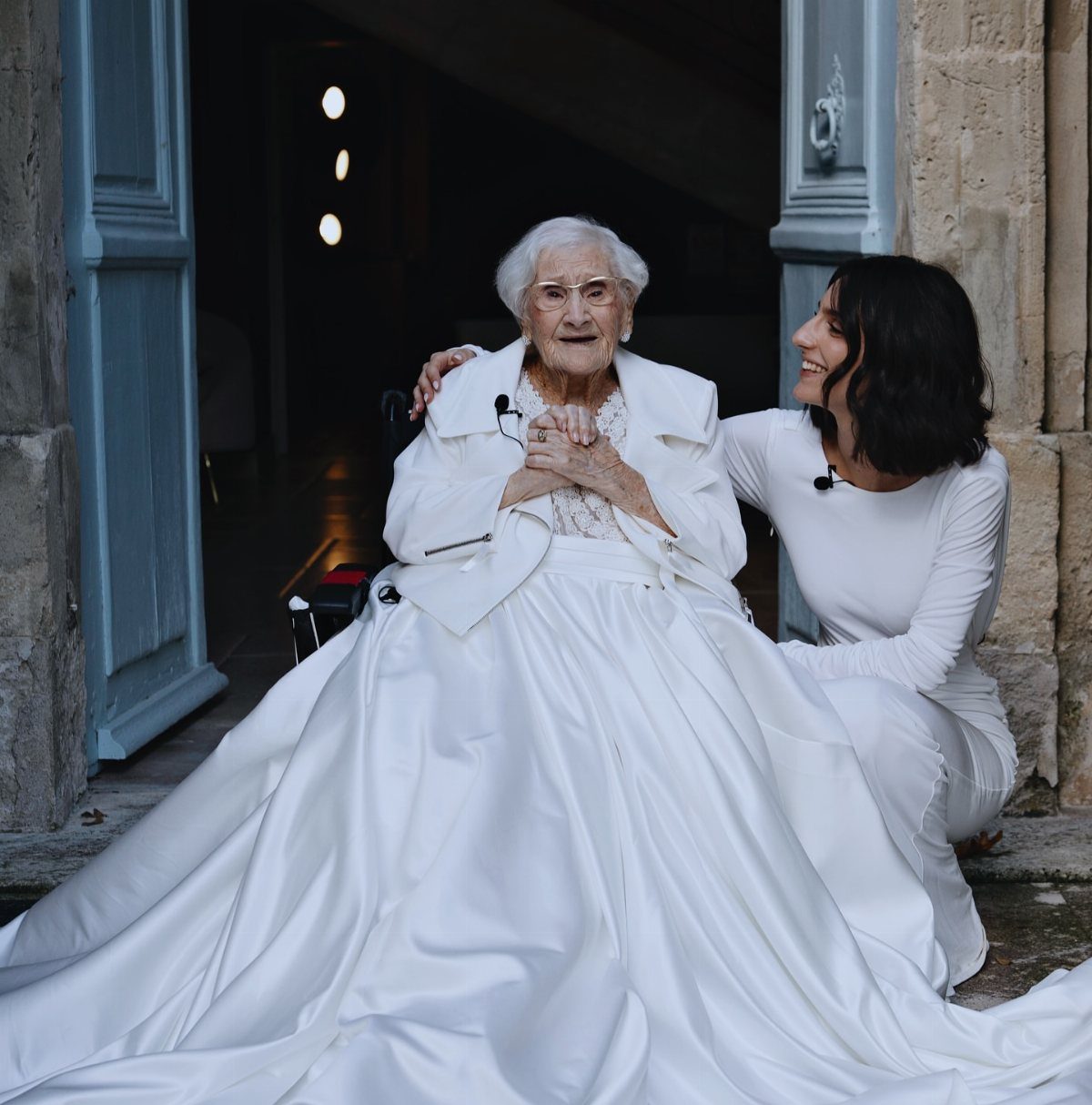 Elderly woman in a voluminous white satin wedding gown and matching short jacket.