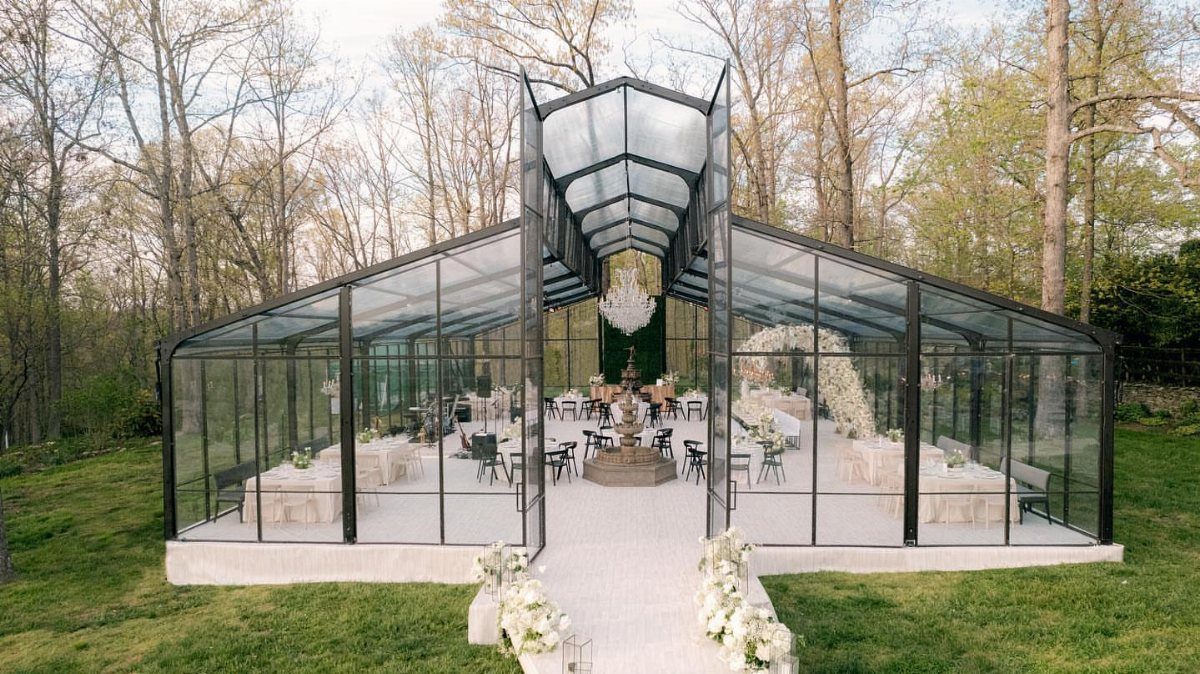 Modern glass greenhouse wedding venue with white tables and chairs in a forest