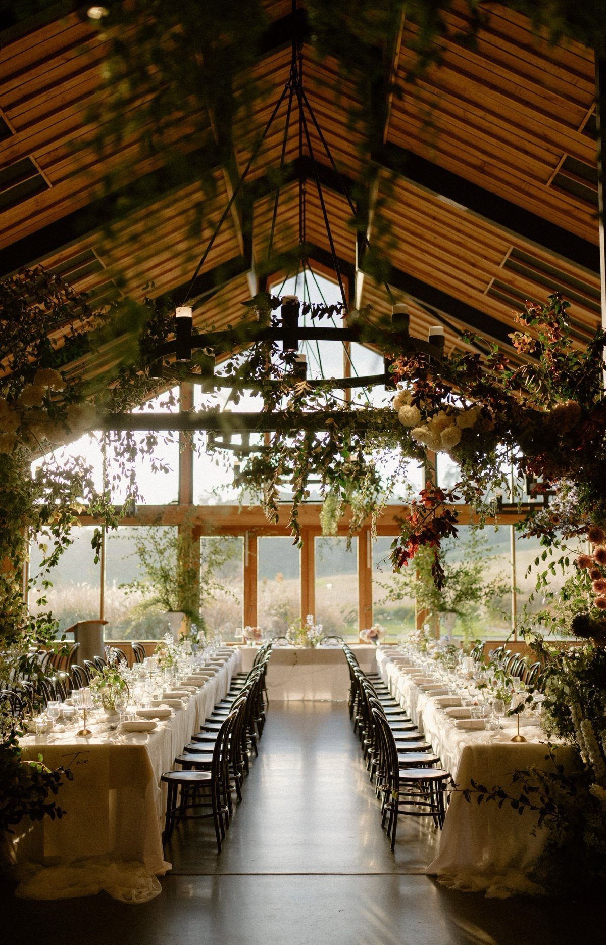 Wedding banquet table set inside a glass greenhouse with wooden beams and greenery.