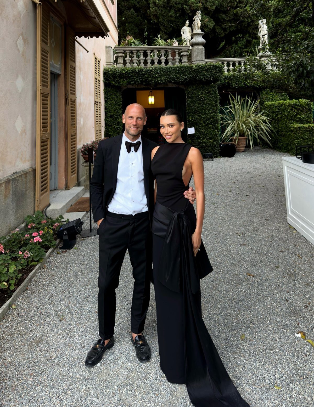 Man in black tuxedo and woman in black gown posing on gravel path.